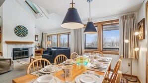 A dining area with a wooden table set for four, a fireplace, and a view of a snowy landscape through large windows.