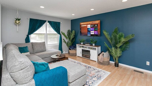 A modern living room with a grey sectional sofa, teal cushions, a small coffee table, and a TV mounted on the wall. There are potted plants and a white cabinet with decorative items. - Quaint 4bd Home - 3 min to Adventureland & Casino! (Altoona)