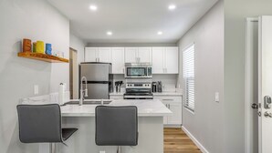 A modern kitchen with white cabinets, a stainless steel refrigerator, a microwave, and an oven. There are two bar stools at the island. - Updated 1 bedroom in paradise! (Waikoloa Village)