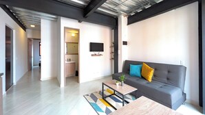 Un salon moderne avec un canapé gris, une table basse en bois et un tapis coloré. Un téléviseur est fixé au mur, et une petite plante est posée sur la table basse.