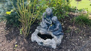 En trädgård med en staty som föreställer två figurer, ett fågelbad och olika växter.