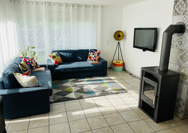 Un salon avec un canapé, des coussins colorés, un tapis à motifs, un téléviseur et un poêle à bois.