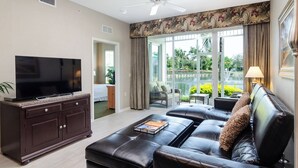 A modern living room with a flat-screen TV, dark wood furniture, a leather sofa, and a view of a garden and water feature through large glass doors. - GARDENIA at Greenlinks - 3 Bedroom Lake View Golf Villa (Naples)