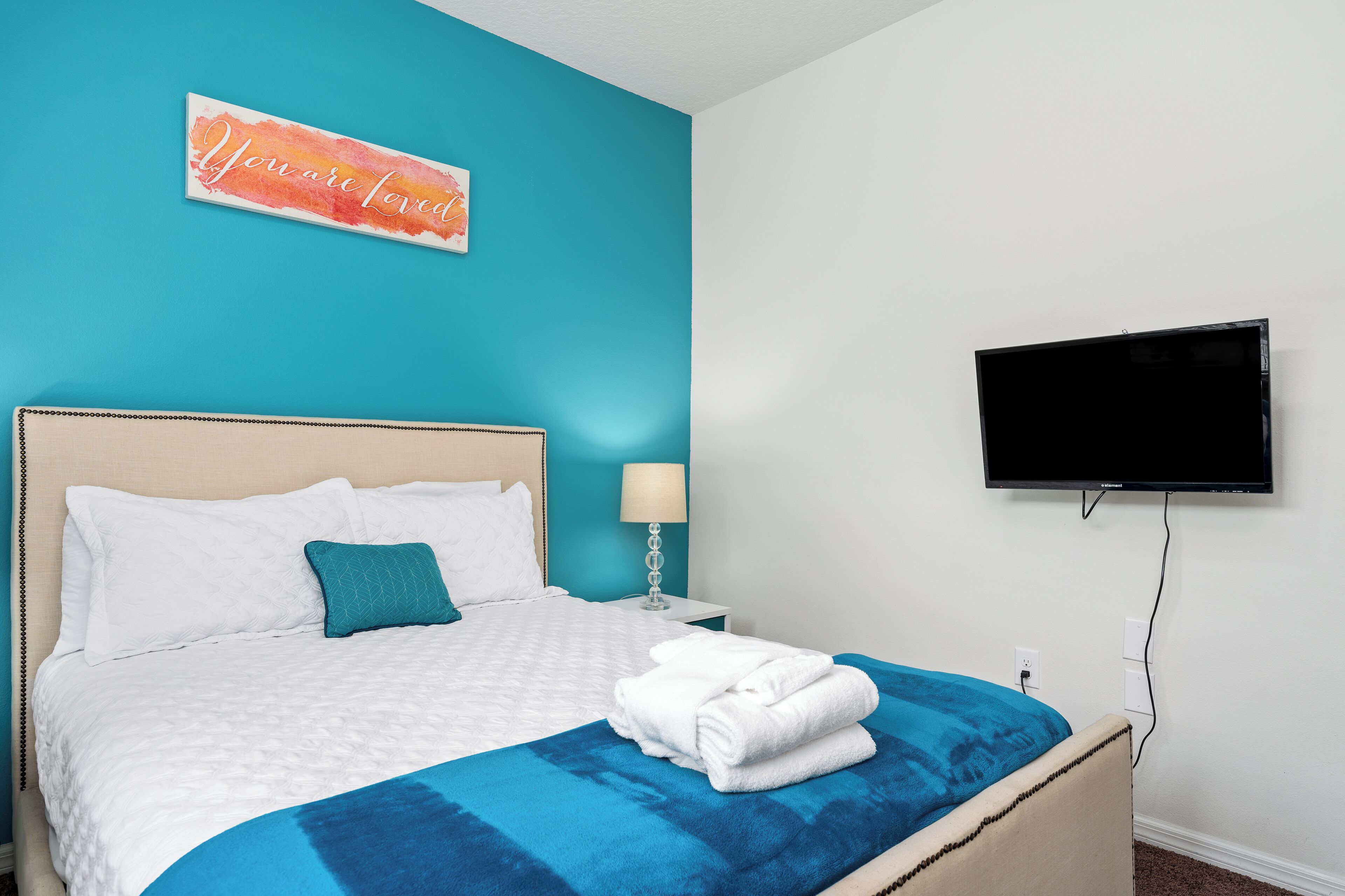 Uma cama bem arrumada com um cobertor azul e almofadas brancas, um abajur e uma televisão de tela plana fixada na parede.