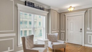 A room with two upholstered chairs, a white dresser, a large window with blinds, a white door, and a ceiling light fixture.