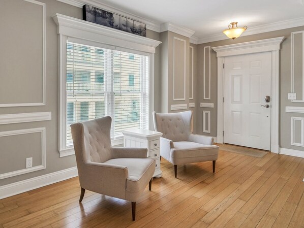 Una habitación con dos sillones tapizados, un aparador blanco, un amplio ventanal con persianas, una puerta blanca y una lámpara en el techo.
