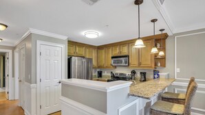 A kitchen with white cabinetry, a stainless steel refrigerator, a microwave, and a stove. There are two pendant lights and a woven chair.