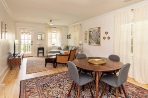 Uma área de refeições com uma mesa redonda de madeira, cadeiras estofadas em cinza, um tapete estampado e um sofá com almofadas.