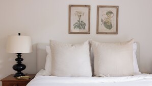 Uma cama com roupa de cama branca, uma mesa de cabeceira com abajur e duas gravuras botânicas emolduradas na parede.