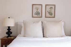 A bed with white bedding, a nightstand with a lamp, and two framed botanical prints on the wall.