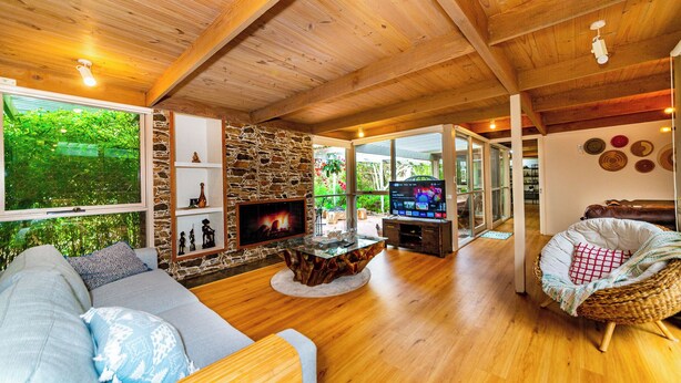 A cozy living room with a stone fireplace, wooden ceiling, and a large window overlooking greenery.