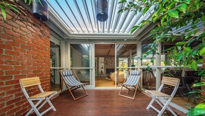 A wooden deck with two striped lounge chairs, a brick wall, and a glass door leading to an interior space.