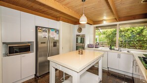 A modern kitchen with white cabinets, stainless steel appliances, a wooden ceiling, and a window overlooking greenery.