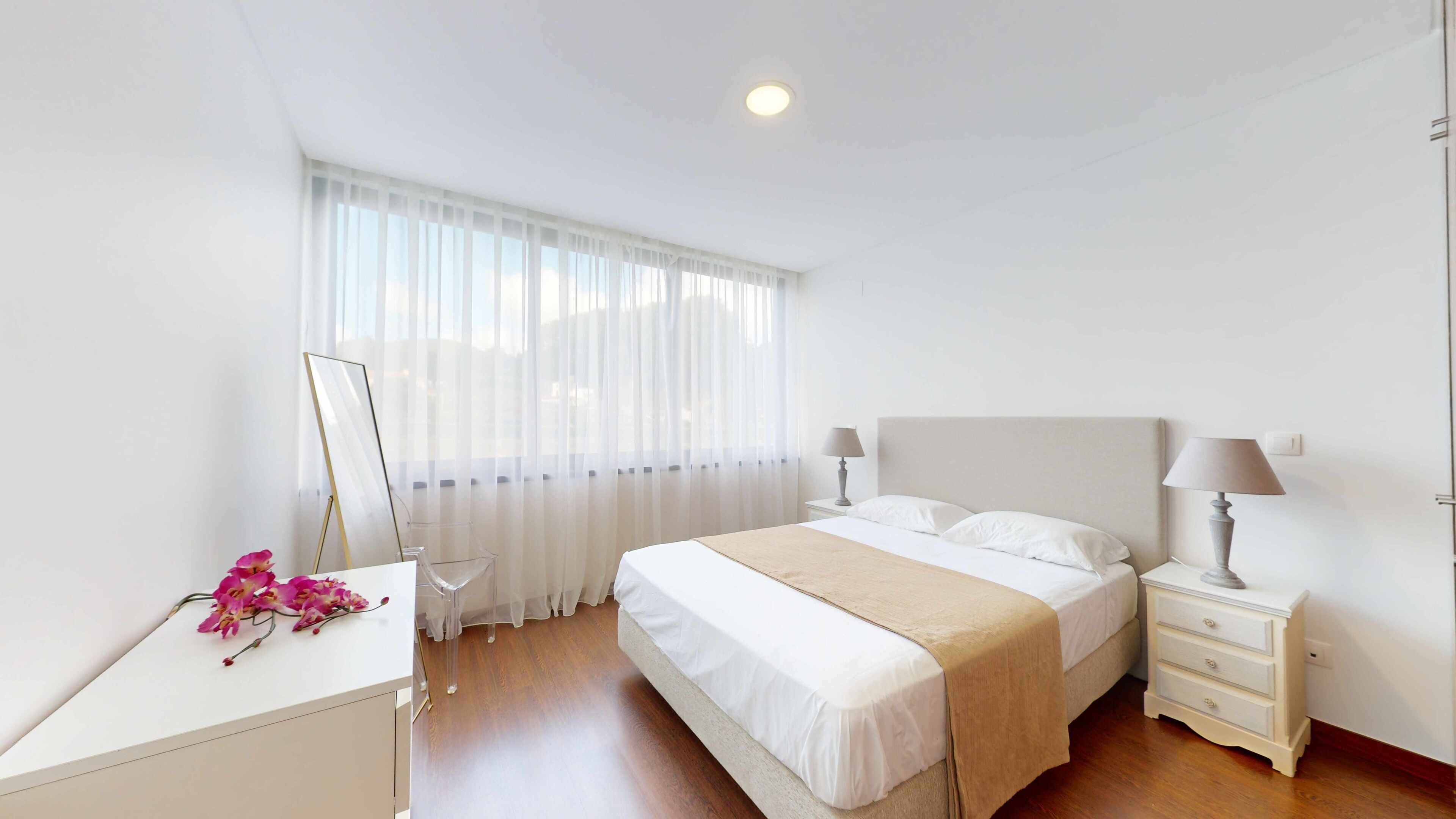 A bedroom with a bed, bedside tables, a desk, and a window with sheer curtains.