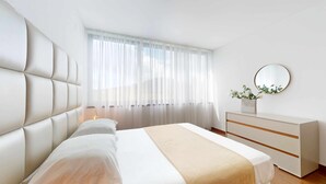 Ein modernes Schlafzimmer mit einer gepolsterten Kopfteil, einem großen Fenster mit durchscheinenden Vorhängen, einem Nachttisch mit Lampe und einer Kommode mit Spiegel.