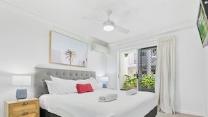 A neatly arranged bedroom with a large bed, a bedside table with a lamp, a ceiling fan, and a view of greenery through a sliding door.