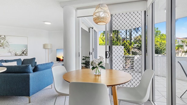 A modern living room with a blue sofa, a wooden dining table, and white chairs. There is a TV and a lamp in the room.