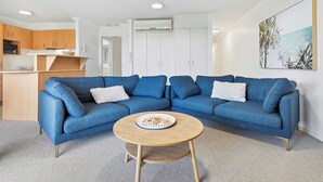 A modern living room with a blue sectional sofa, a round wooden coffee table, and a kitchen area in the background.