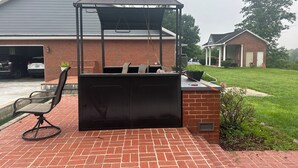 An outdoor grill area with a brick patio, a chair, and a house in the background. - Large private room in a large home nestled in the mountain of Johnson county Ky. (Hagerhill)
