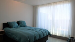 A bedroom with a bed, a nightstand, a window with sheer curtains, and a view of the outdoors.