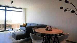 A modern living room with a round wooden table, grey upholstered chairs, a grey sofa, and a large window offering a view.