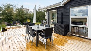 Eine Holzterrasse mit Essbereich, Gartenmöbeln und einem großen Fenster mit Blick auf einen Garten.