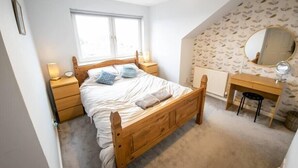 A bedroom with a wooden bed, a bedside table with a lamp, a dresser with a mirror, and a window with a view. - Tranquil 2Bed/2Bath Duplex Falkirk (Falkirk)