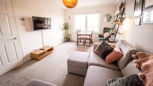 A modern living room with a grey sofa, a flat-screen TV mounted on the wall, a dining area with a table and chairs, and a large window providing natural light. - Tranquil 2Bed/2Bath Duplex Falkirk (Falkirk)