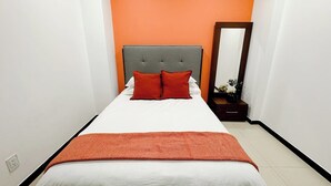 Cama bem arrumada com cabeceira cinzenta, roupa de cama branca e duas almofadas vermelhas.