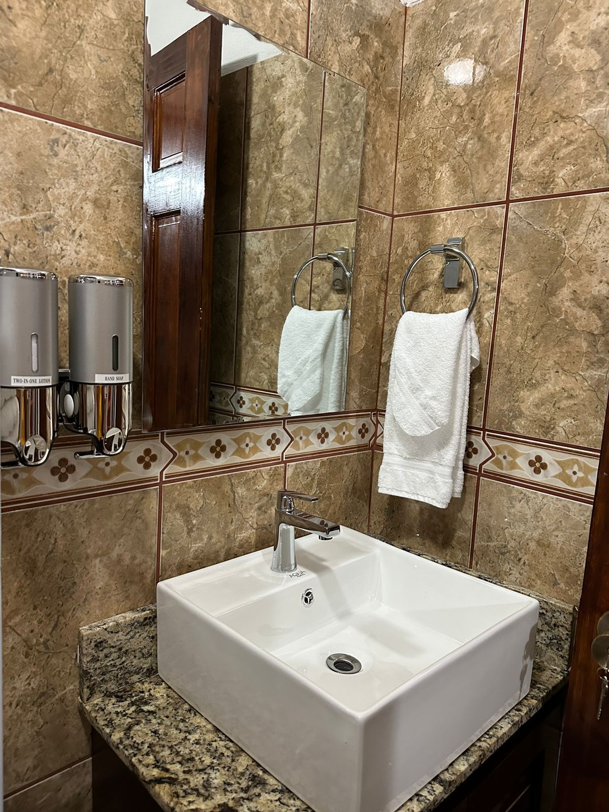 A bathroom with a white sink, marble tiles, a mirror, and two hand soap dispensers.