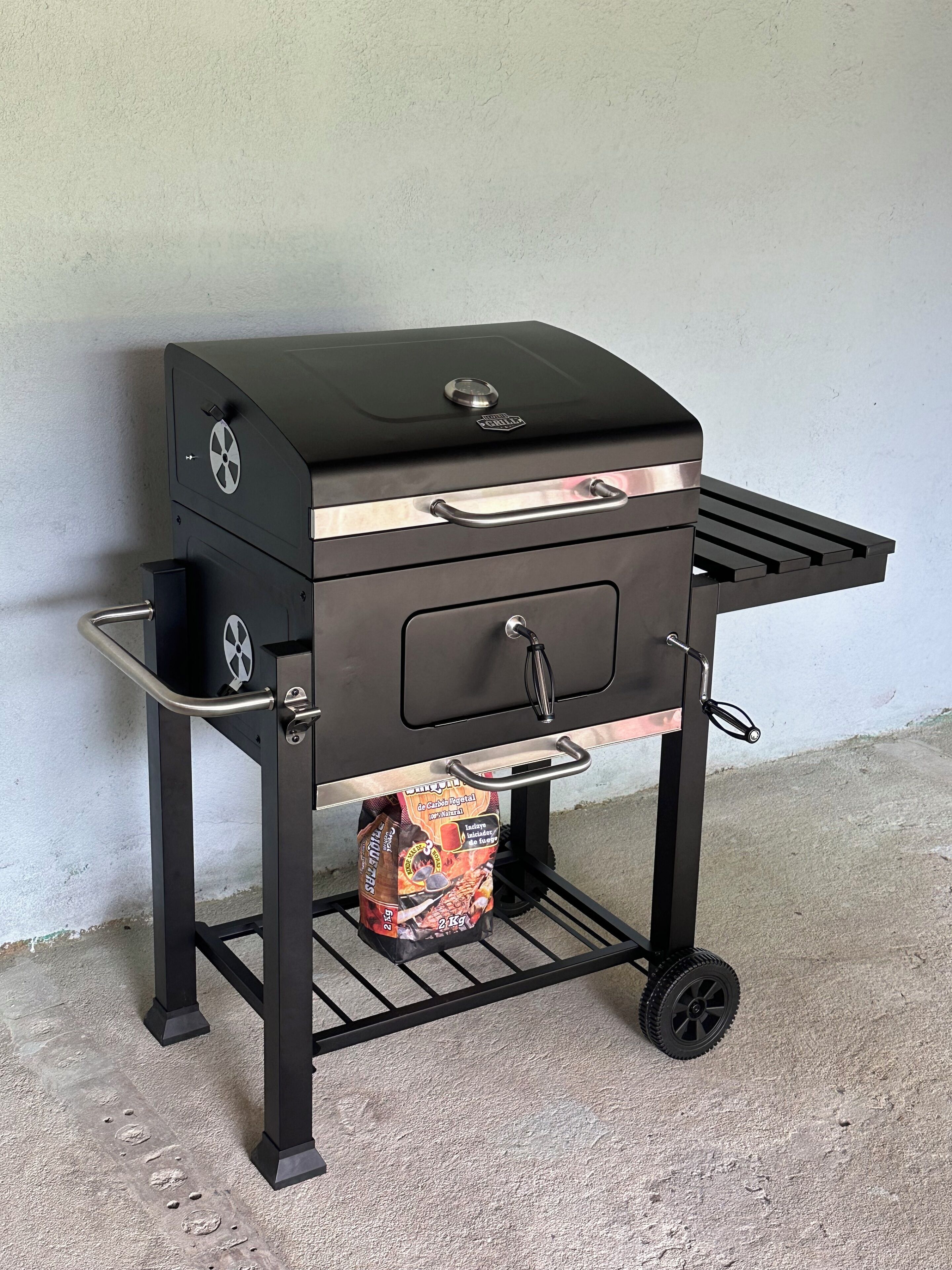 A portable barbecue grill with wheels, a side shelf, and a bag of charcoal underneath.