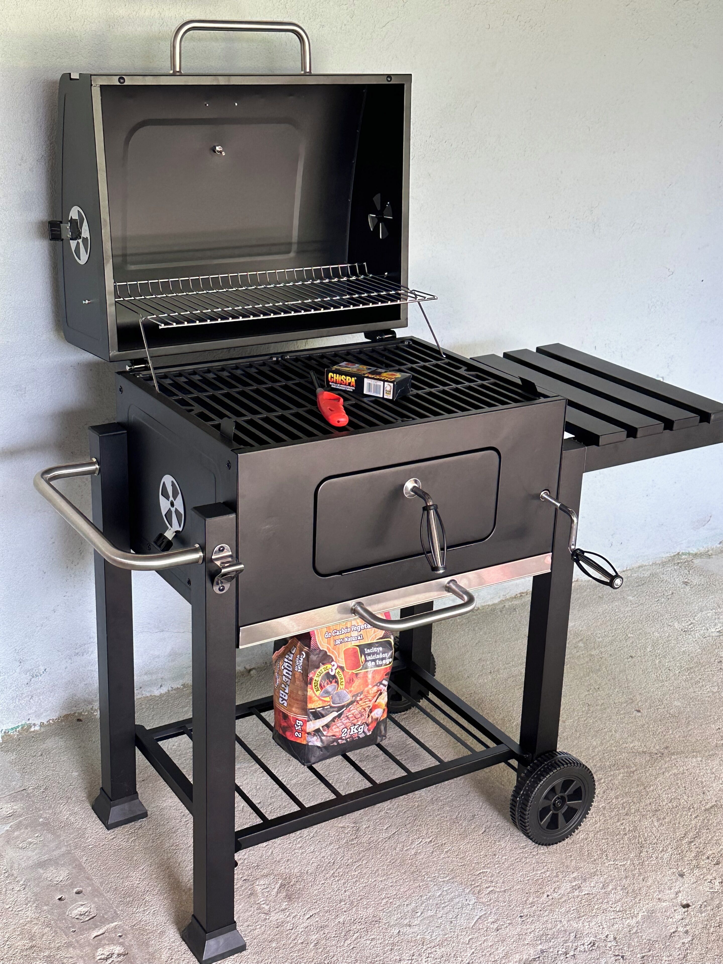 Una parrilla portátil con tapa, asa y rueda para facilitar su transporte.
