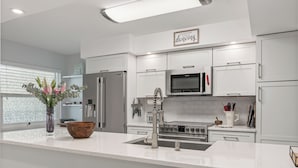 A modern kitchen with white cabinetry, stainless steel appliances, and a central island with a sink and a vase of flowers. - Amazing water/sunset views, luxury 2BR/2BA PENTHOUSE 9 mi from Anna Maria beach. (Bradenon)