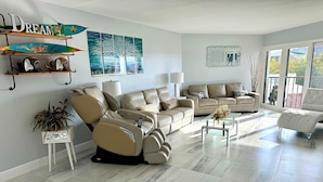 A modern living room with a beige sofa, a recliner, and a glass coffee table. There are decorative items on shelves and a painting on the wall. - Amazing water/sunset views, luxury 2BR/2BA PENTHOUSE 9 mi from Anna Maria beach. (Bradenon)