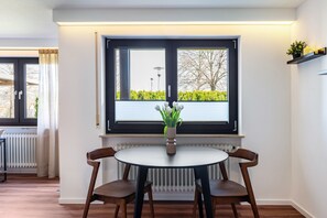 Un coin repas comprenant une table ronde, deux chaises, un vase avec des fleurs, une fenêtre avec des stores et un radiateur.
