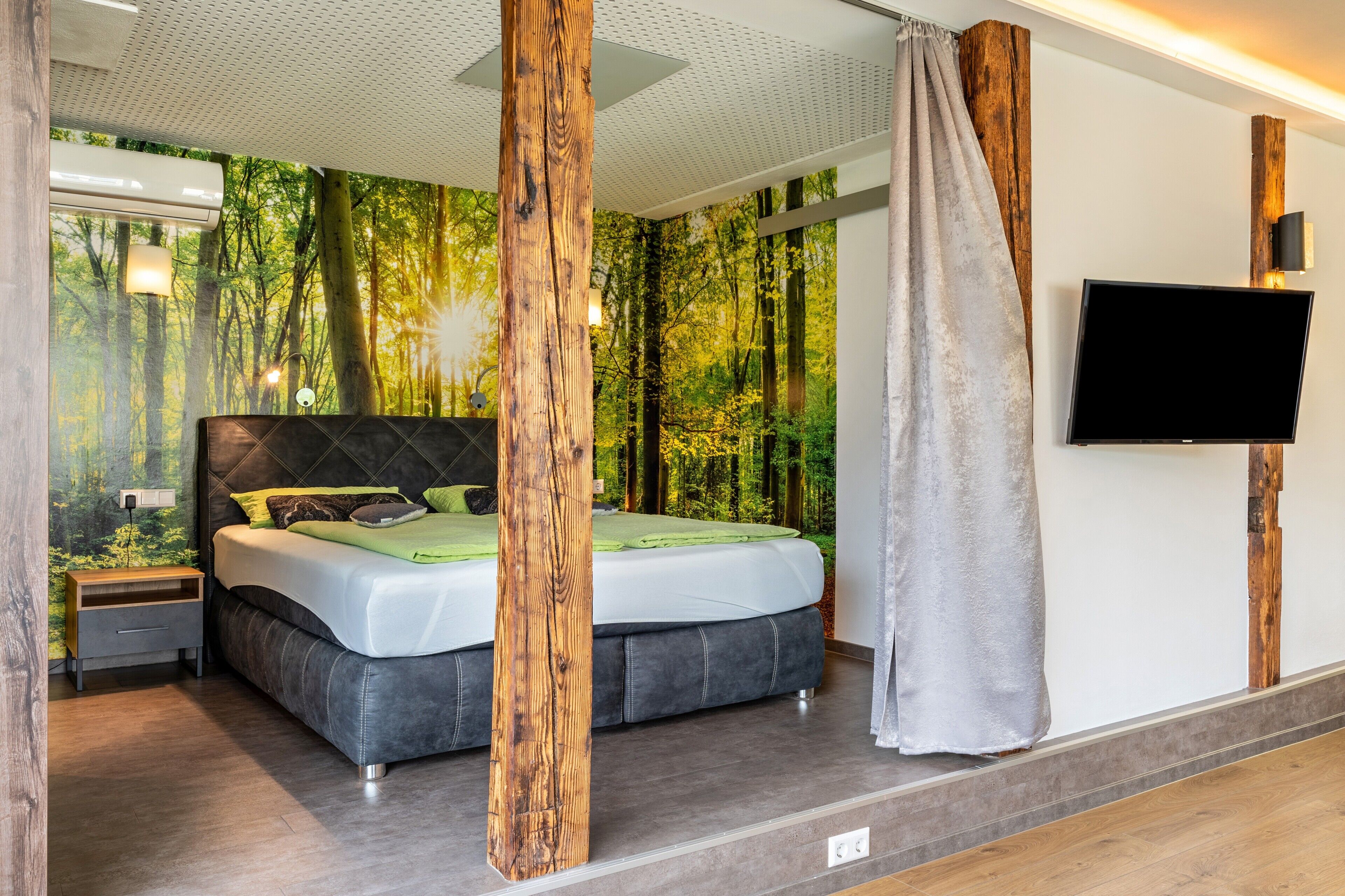 A modern bedroom with a large bed, a TV, and a forest-themed wall mural.