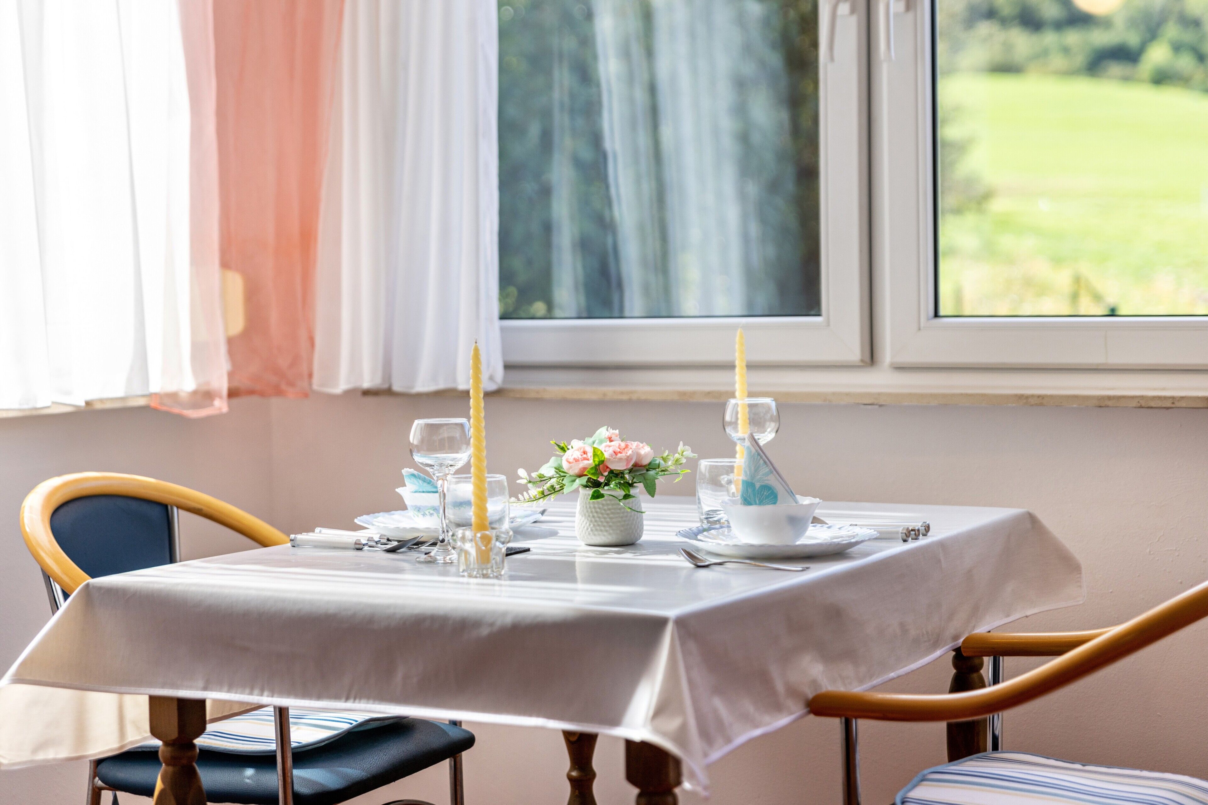 A dining table with a white tablecloth, two candles, a vase with flowers, and a window with a view of a green field.