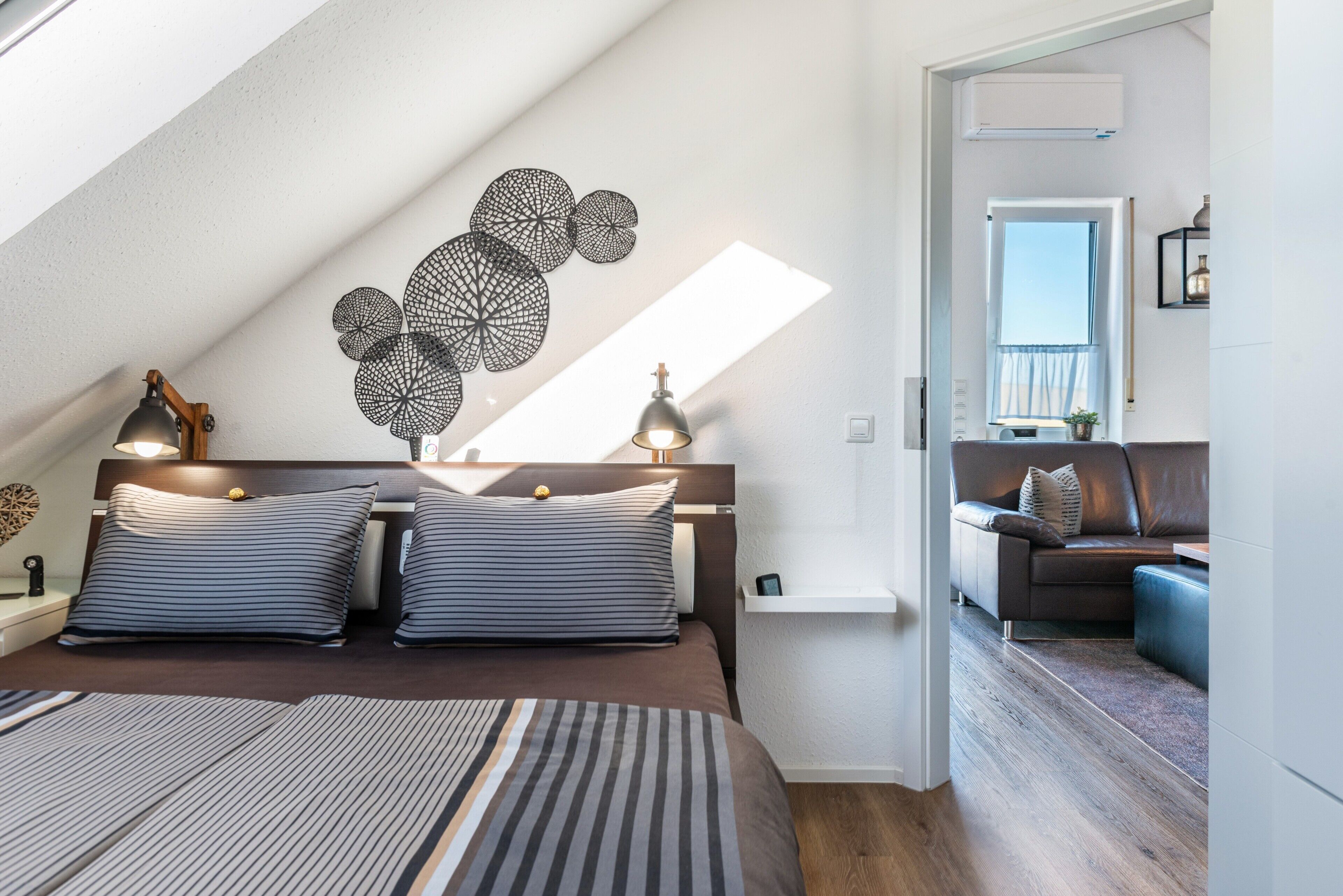 Quarto moderno com teto inclinado, uma cama grande com roupa de cama listrada, duas luminárias de cabeceira e uma obra de arte na parede.