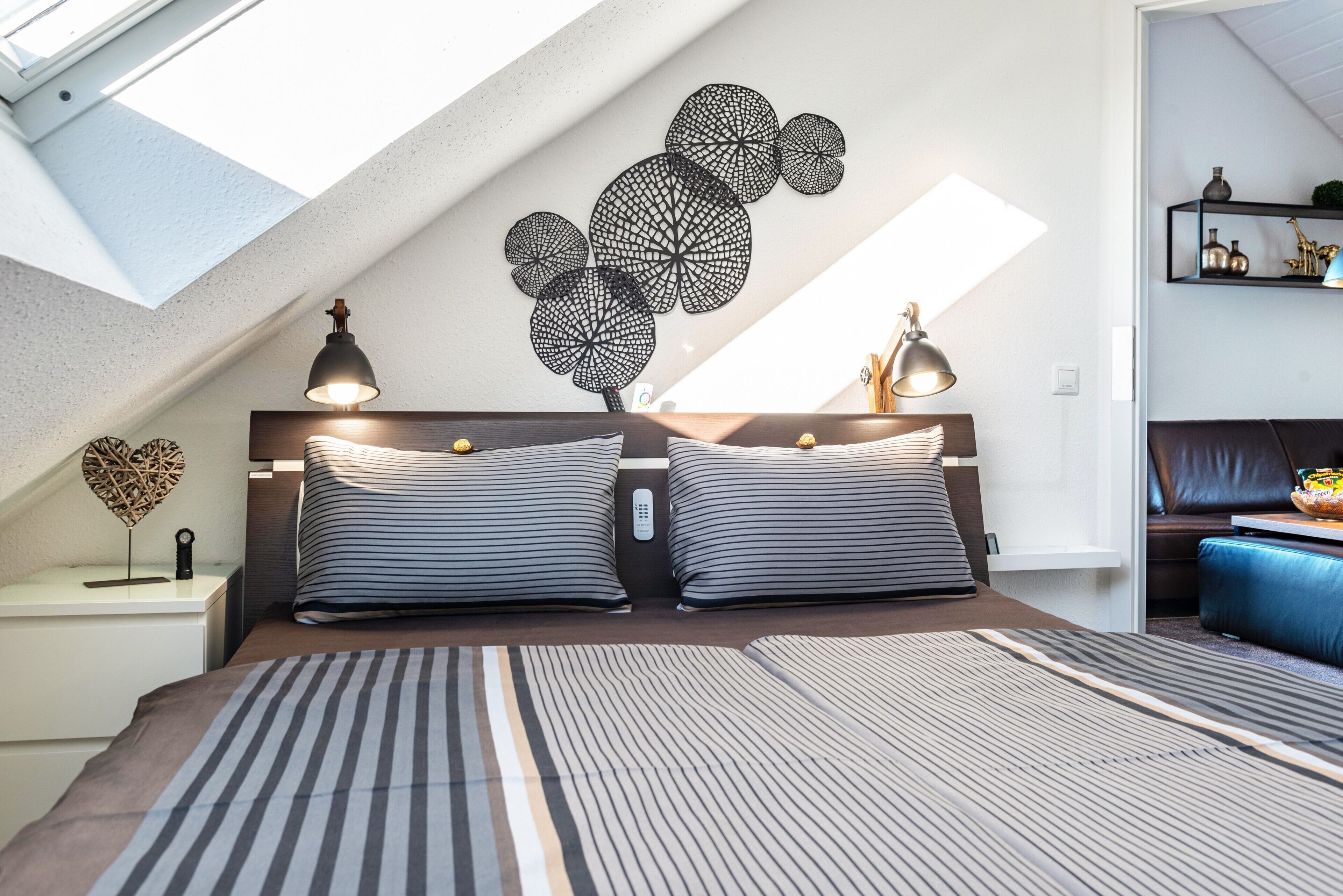 A bedroom with a skylight, a bed with striped bedding, a wall art piece, and a bedside table with lamps.