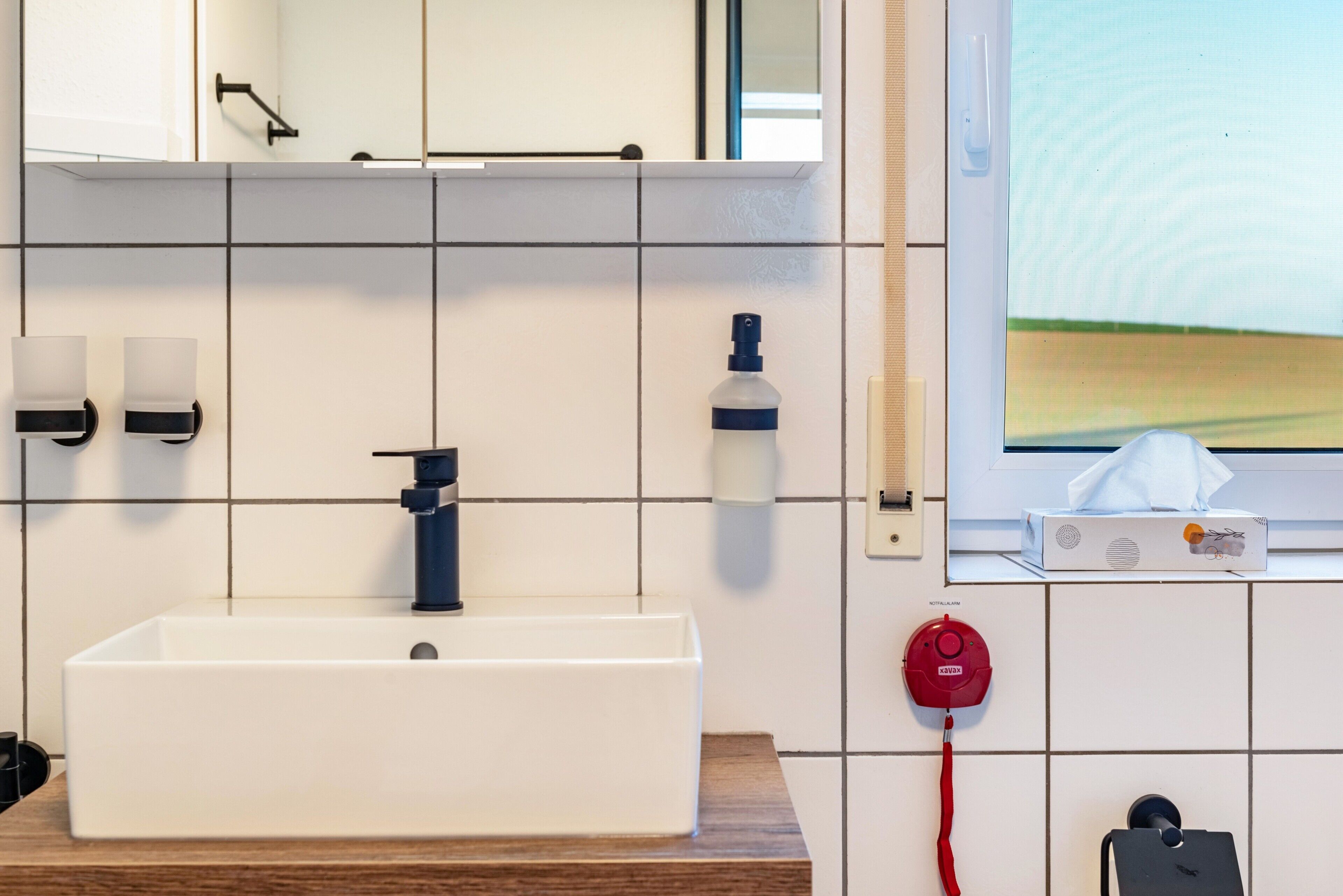 A bathroom with a white sink, a mirror, a hand dryer, a soap dispenser, and a tissue box on the wall.
