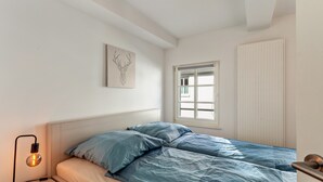 Une chambre à coucher comprenant un lit, une lampe de chevet, une fenêtre, un radiateur et une œuvre d’art accrochée au mur.