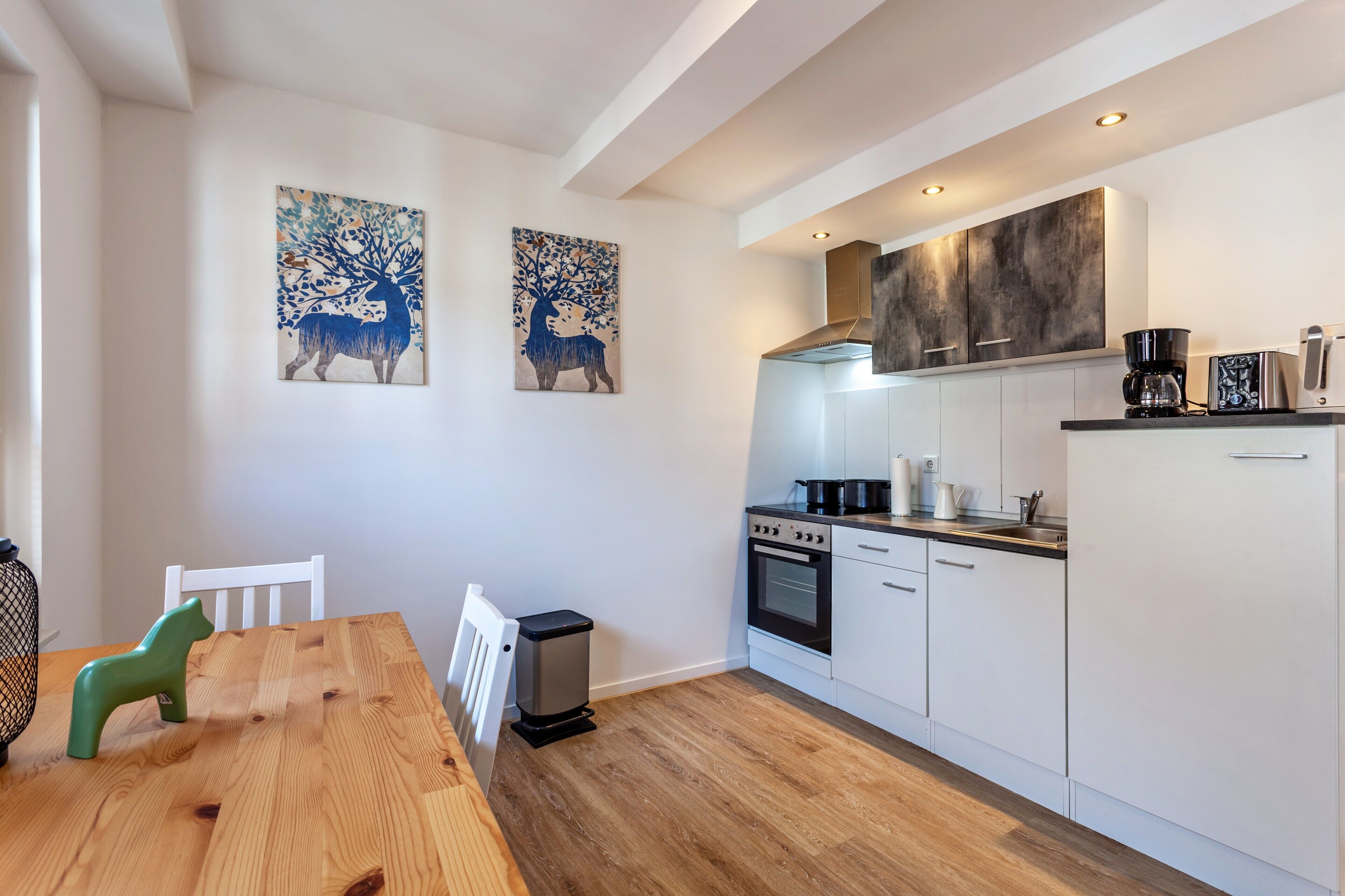 Een moderne keuken met witte kastjes, een fornuis en een koffiezetapparaat. Er hangen twee schilderijen van herten aan de muur.