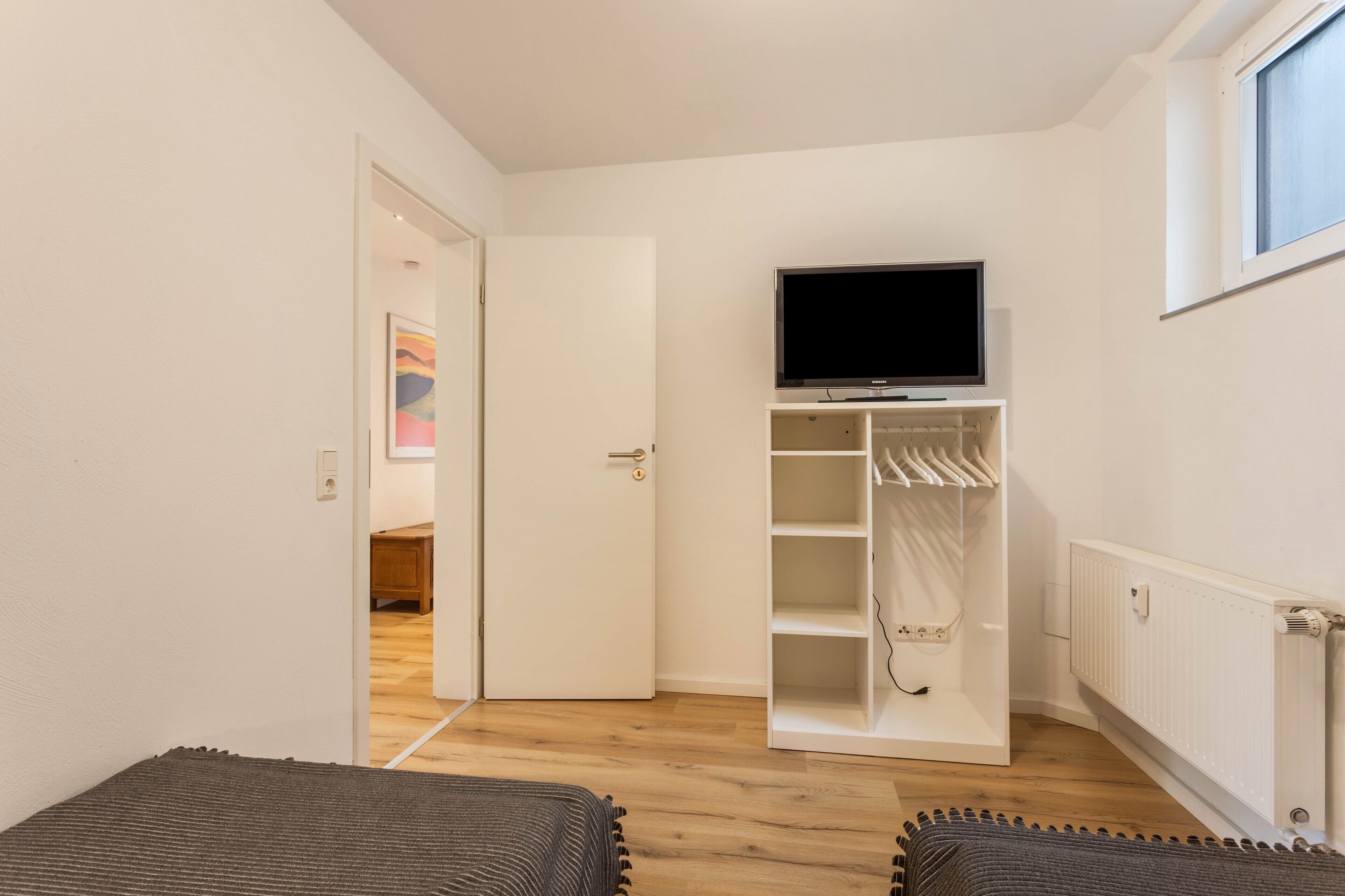 Ein Zimmer mit Fernseher auf einem Schrank, einem weißen Kleiderschrank, einem Heizkörper und einer Tür zu einem anderen Raum.