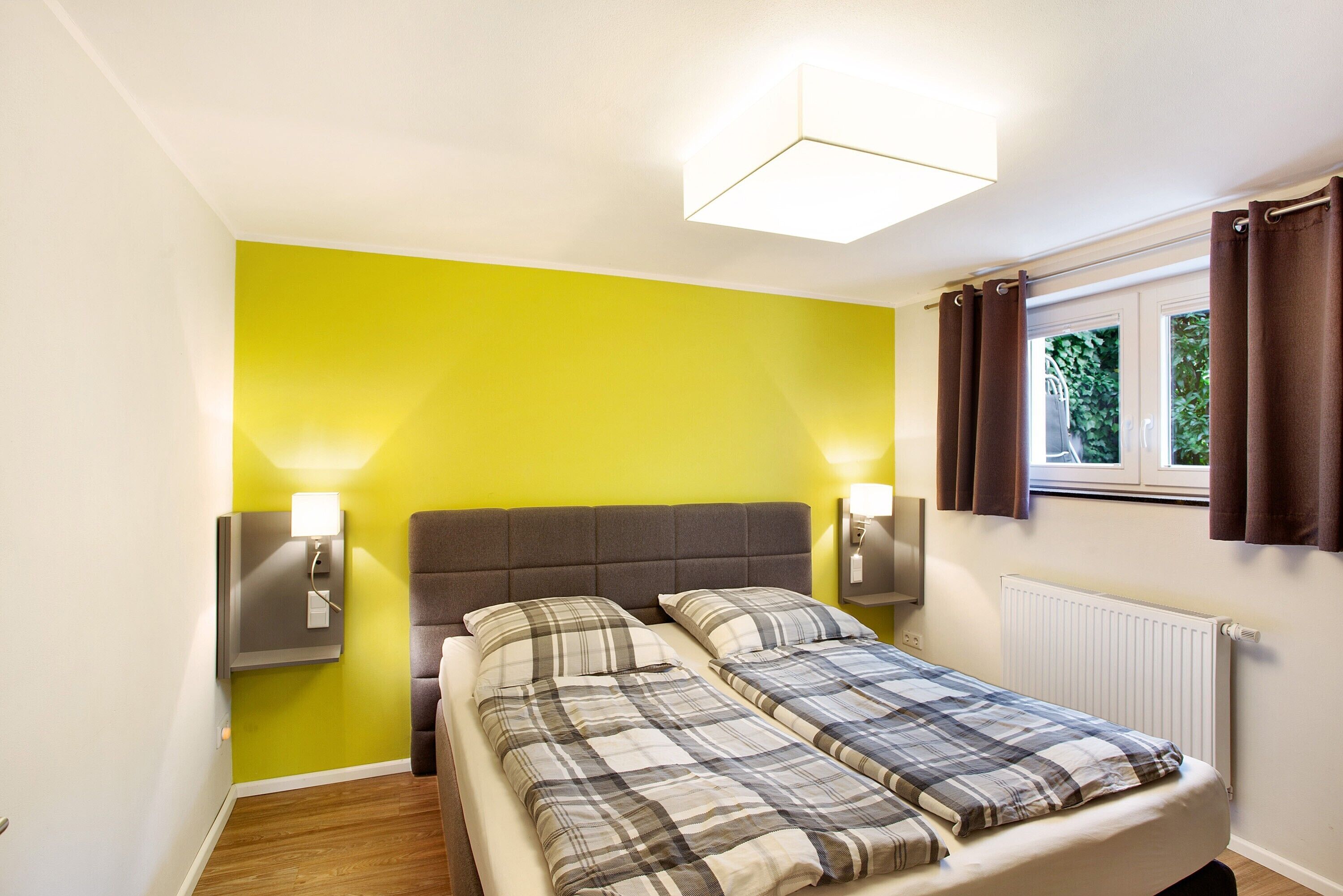 Une chambre avec un lit, deux lampes de chevet, une fenêtre avec des rideaux et un luminaire fixé au mur.