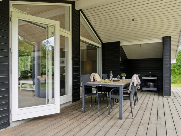 A covered outdoor patio with a wooden table and chairs, a grill, and a glass door leading to a garden.