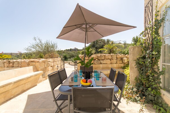 An outdoor dining area with a table, chairs, a large umbrella, and a fruit bowl.