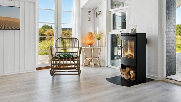 A room with a large window, a wooden floor, a black fireplace, and a wicker chair. - "Blaguna" - 600m from the sea by Interhome (Thisted)