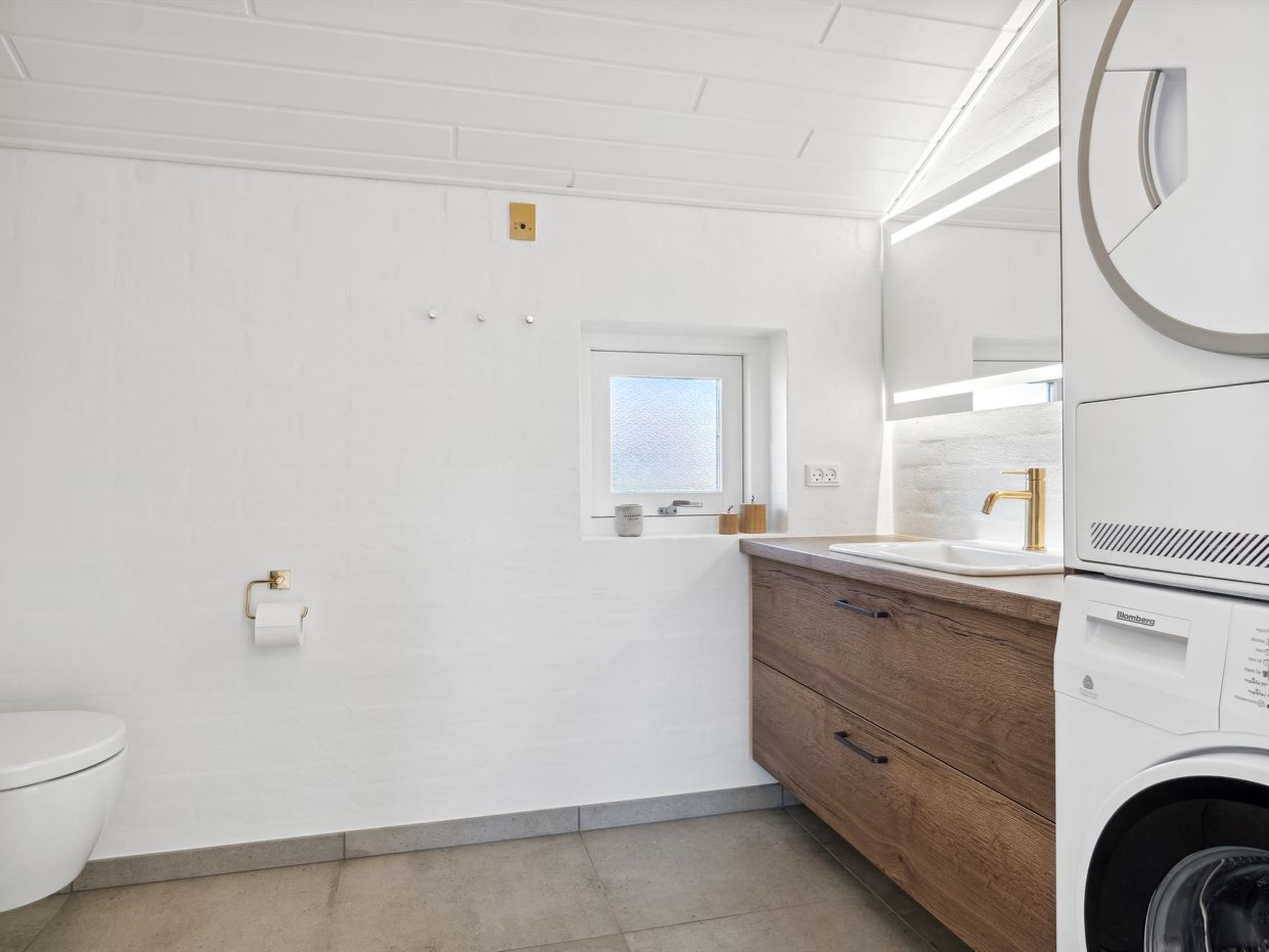 Une salle de bain moderne avec des toilettes blanches, une fenêtre, un meuble-lavabo en bois et un lave-linge.
