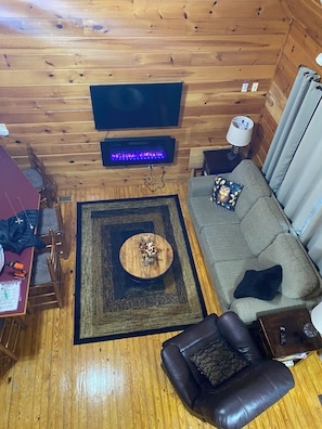 A cozy cabin-style room with a wooden interior, a flat-screen TV mounted on the wall, a fireplace with a blue flame, a grey sofa with decorative pillows, a brown leather chair, and a small wooden table with a lamp.