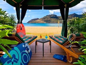 Um espaço de lazer à beira-mar, com duas espreguiçadeiras, uma mesa com bebidas e uma prancha de surf.